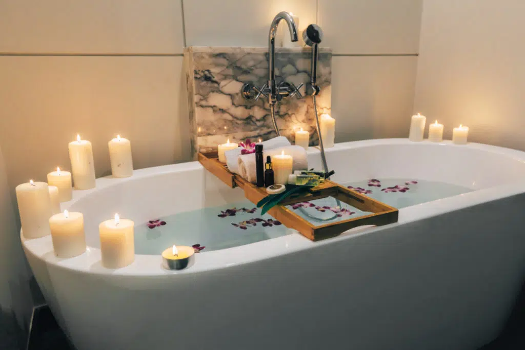 une baignoire blanche est remplie d'eau avec des fleurs qui flottent. des bougies allumées sont disposées partout autour du bord de la baignoire. un support en bois est posé sur la baignoire avec des bougies, serviettes et autres objets détente.