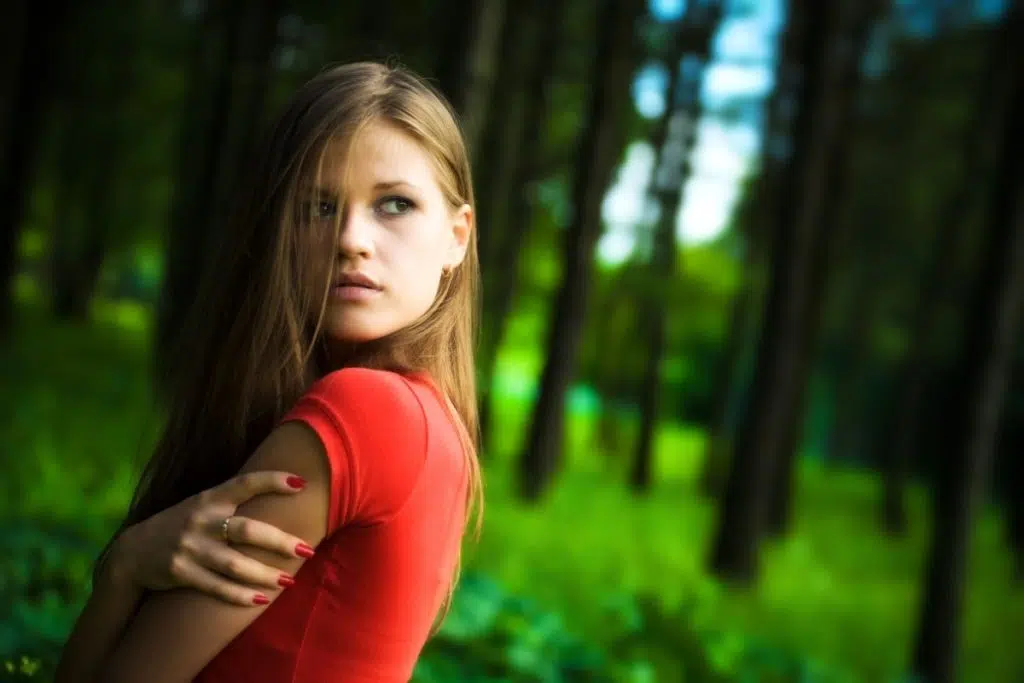 dans une forêt dense, une femme de profil en t-shirt rouge regarde derrière elle la tête tournée vers l'arrière. elle a les bras croisés.