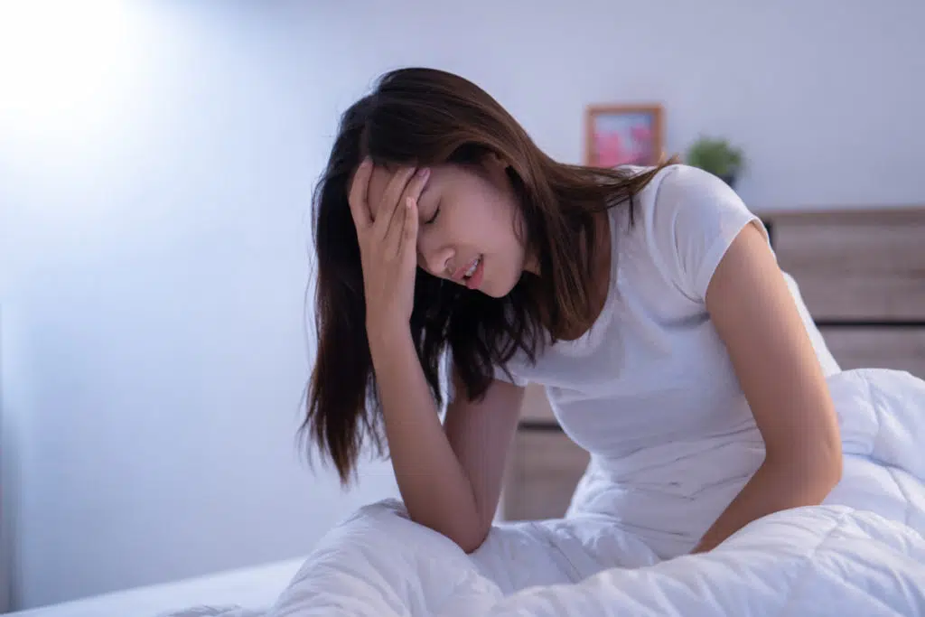 une femme assise sur un lit en tee-shirt blanc, se tient le front, tête baissée. Elle n'a pas l'air d'aller bien