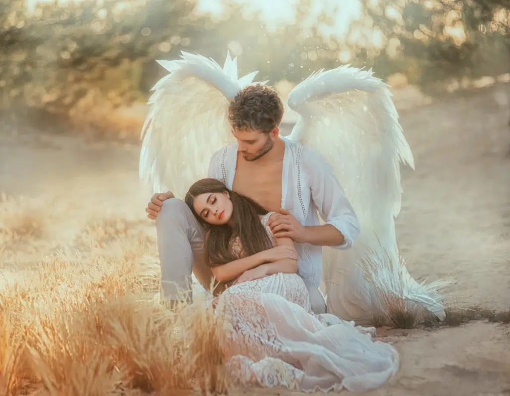 un ange avec de grands ailes blanche a une jeune femme les yeux fermés, posée sur son genou. ils sont dans un champ.