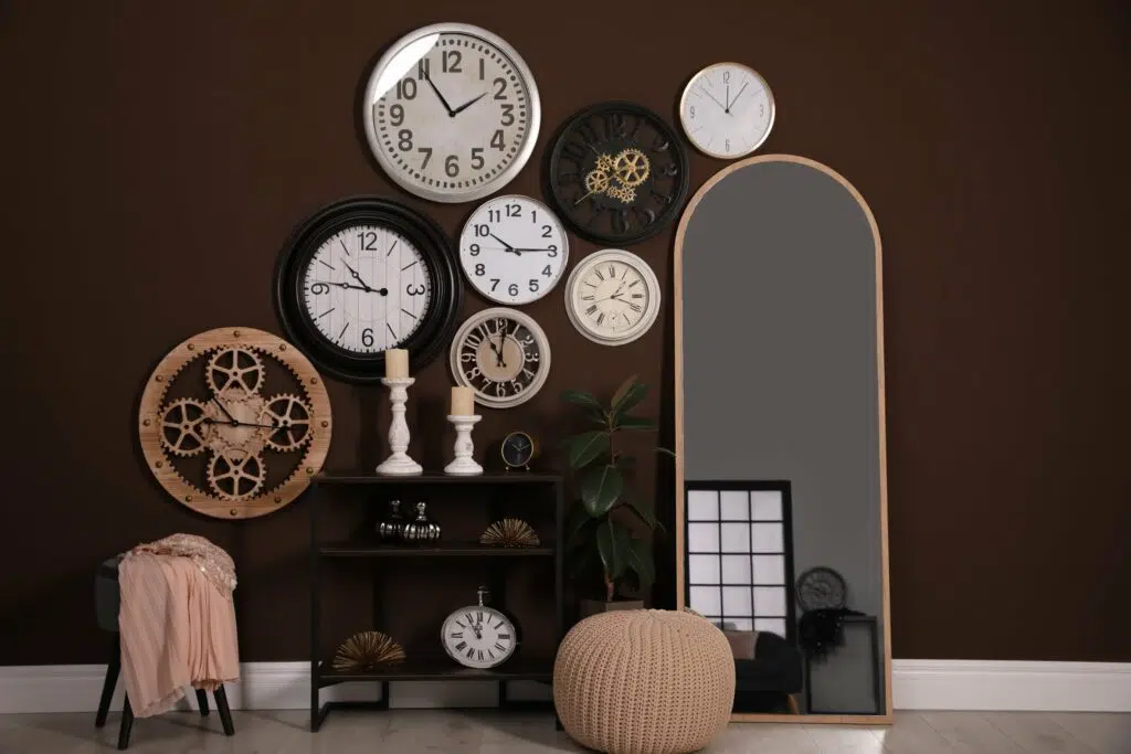 plusieurs horloges accrochées à un mur marron avec un grand miroir, un meuble console, un pouf et un tabouret. 