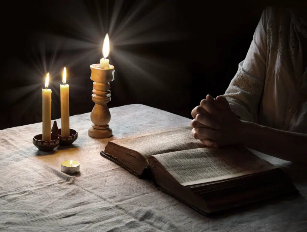 une personne prie avec les mains jointes sur une bible posée sur une table avec trois bougies blanches. 