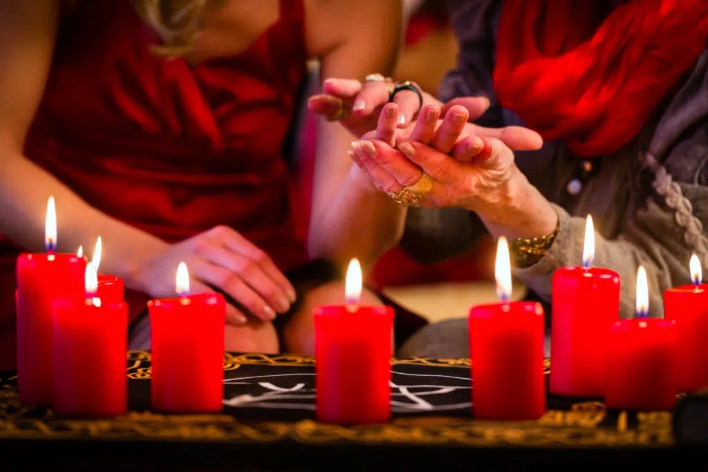 une voyante tient la main d'une femme enrouge au dessus de plusieurs bougies rouges. 