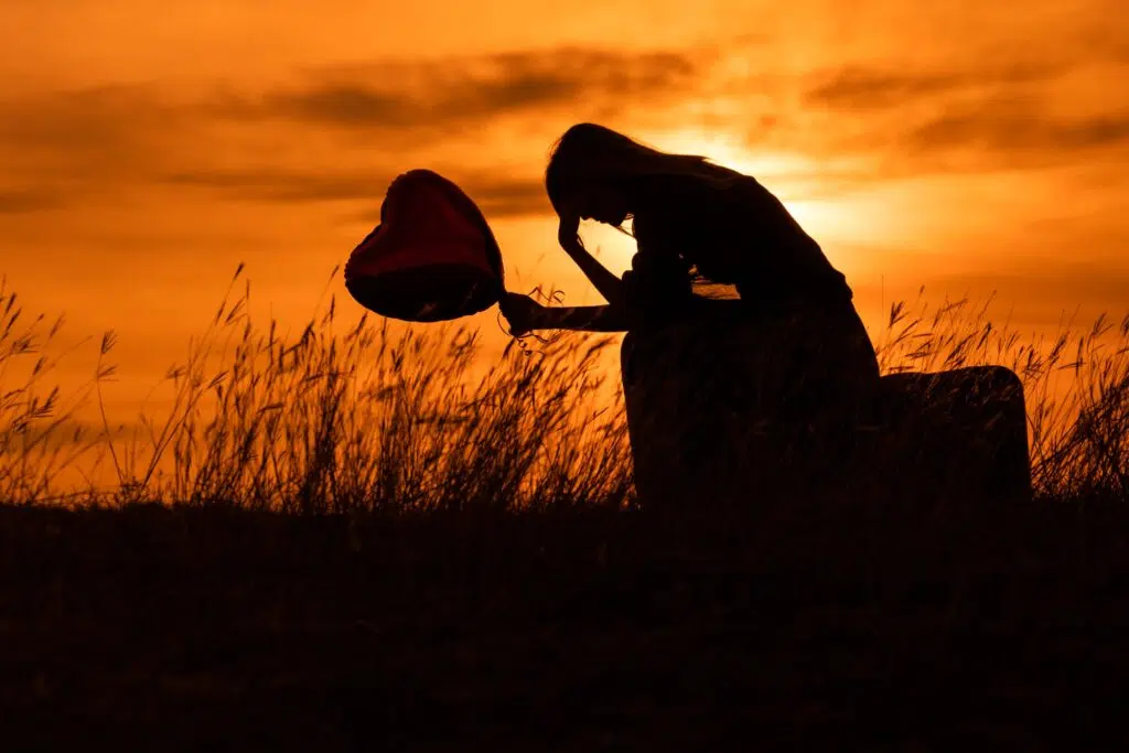 Silhouette d'une femme triste assise, se tenant la tête dans une main et dans l'autre tenant un ballon en forme de cœur. Le tout sur un fond de coucher du soleil.