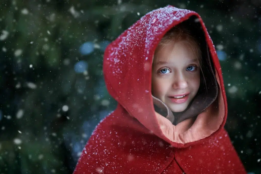 Jeune fille aux yeux bleus avec un sourire timide, vêtue d'un manteau rouge à capuche dehors sous une fine neige