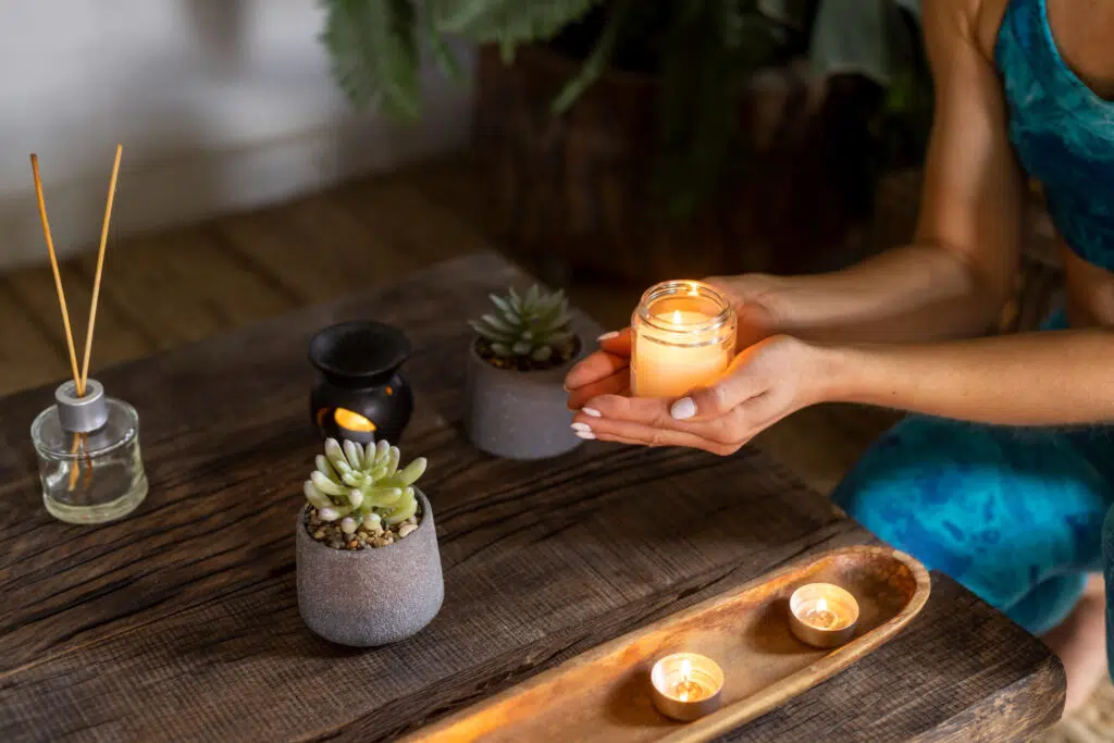 mains d'une femme en bleu qui tiennent une bougie en coupe au dessus d'une table en bois foncé où sont posés des pots de petites plantes, de l'encens et un bruleur d'encens. 