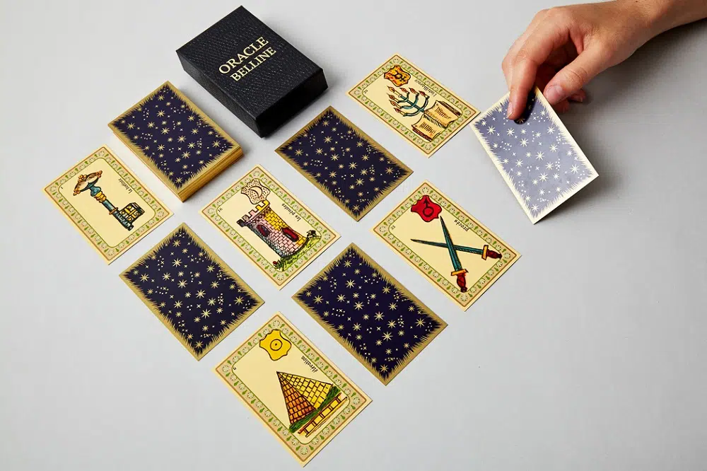 des cartes de jeu et une boite oracle de belline, avec une main qui retourne la carte sur le coin en haut 