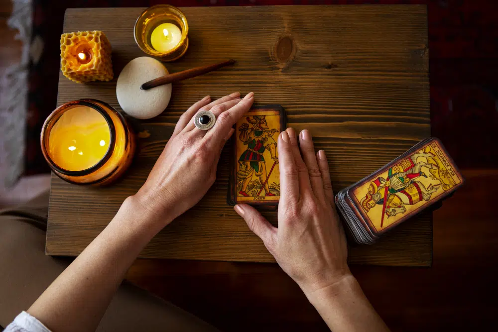 vue de haut des mains d'une femme qui tiennent un jeu de tarot posé sur une table en bois à côté d'un jeu de tarot, des bougies et de l'encens sur les côtés