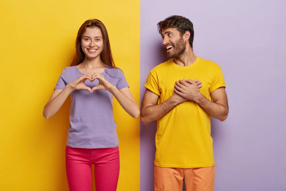 une jeune femme en tshirt violet sur un fond jaune, à côté d'un jeune en tshirt jaune sur un fond violet, qui la regarde 