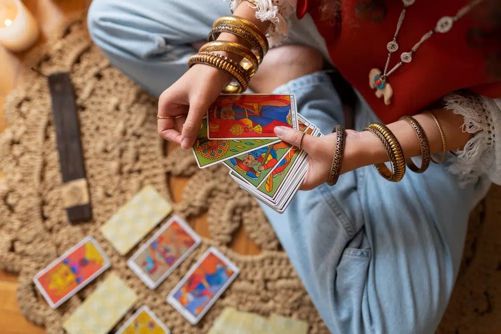 les mains d'une jeune femme en jeans bleu qui tiennent un jeu de tarot  assise sur un tapis couleur sable où sont posées des cartes de tarot